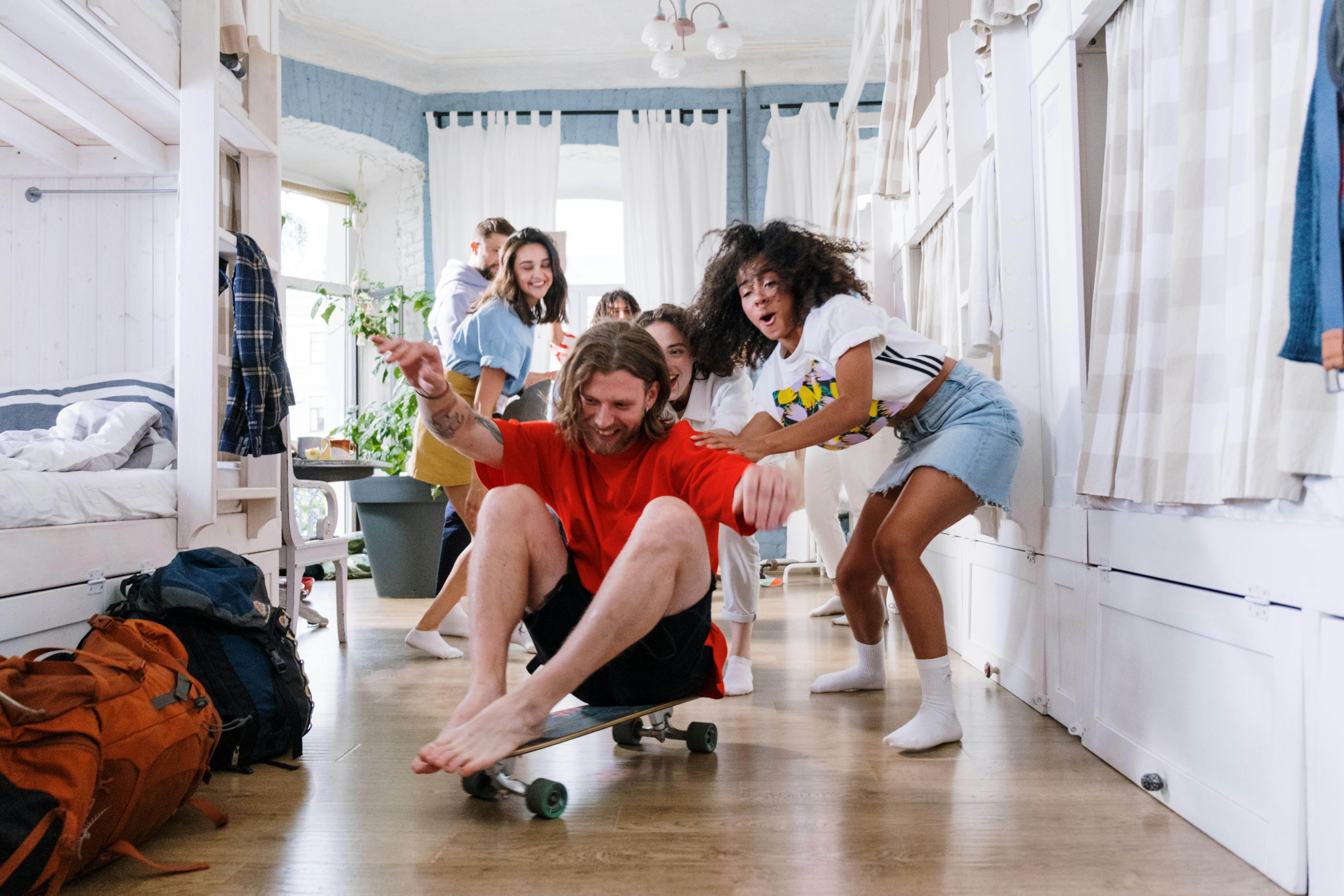 Image of A bunch of young adult friends playing around in a blue-painted room with large windows behind them. A young man is being pushed around on the floor, laughing, on a skateboard.